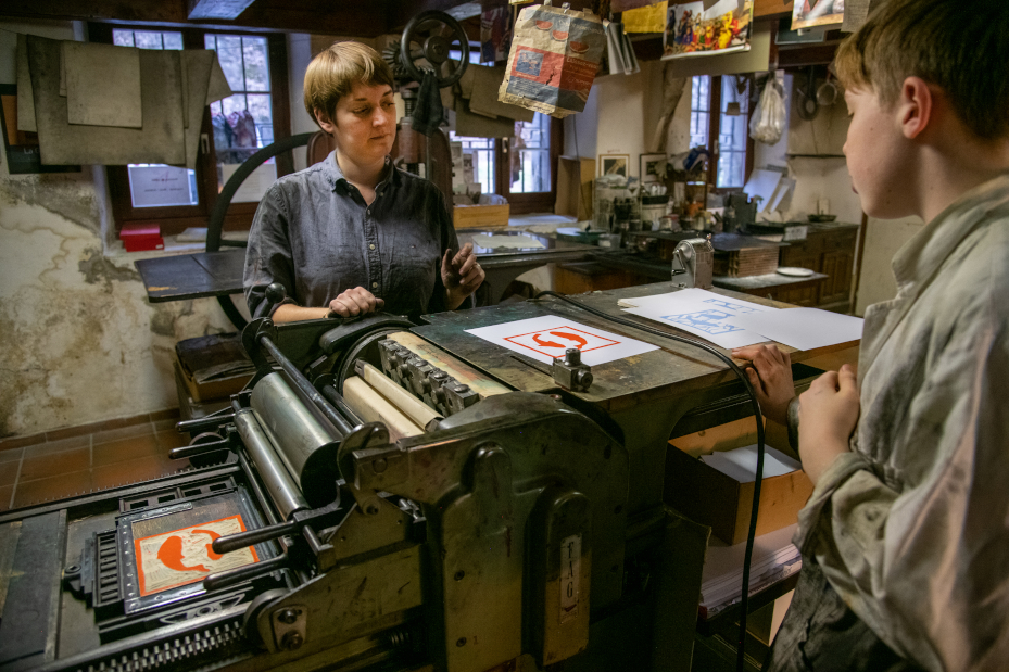 Atelier de gravure au Pilon-à-Os : une animatrice et un jeune participant devant une presse typographique, avec des tirages aux motifs colorés